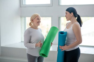 Sarışın kadın ve esmer kadın ellerinde yoga halılarıyla bir şeyler tartışıyorlar.