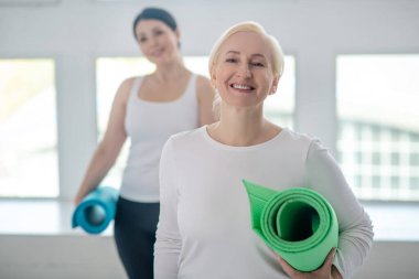 Sarışın kadın yoga halısıyla ayakta duruyor ve gülümsüyor, esmer kadın arkasında duruyor.