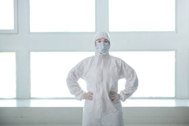 Koruyucu giysiler, tıbbi maske ve gözlük takan bir tıp çalışanı elleri belinde duruyor.