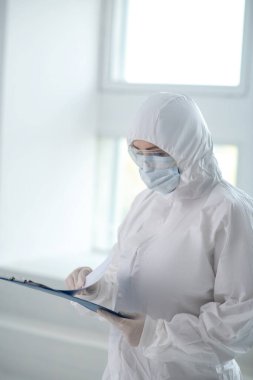 Koruyucu giysiler, tıbbi maske ve gözlük takan bir tıp çalışanı. Şarjör klasöründeki kağıtlara bakıyor.