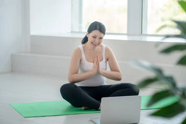 Morena hembra realizando namaste, teniendo en línea yoga lección, sonriendo — Foto de Stock