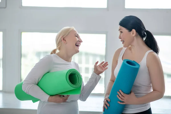 Rubia femenina y morena femenina sosteniendo alfombras de yoga y riendo — Foto de Stock