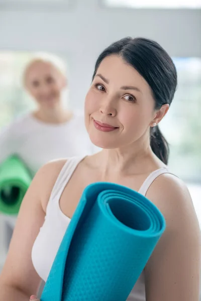 Bonita morena mujer sosteniendo alfombra de yoga y sonriente, rubia mujer de pie en el fondo — Foto de Stock