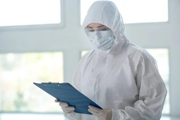 Medical worker in protective clothing, medical mask and goggles checking something on clip folder