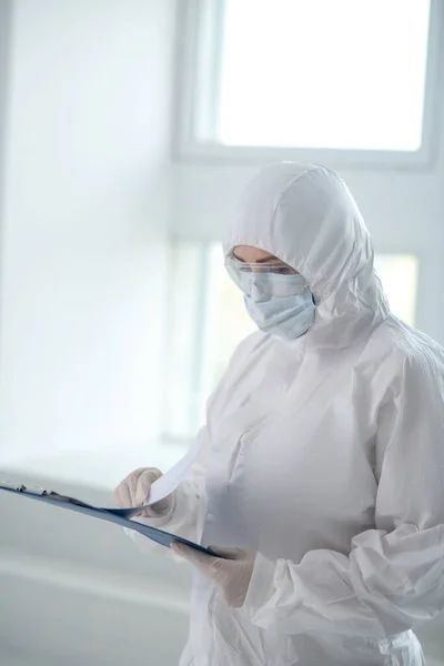 Медичний працівник у захисному одязі, медичній масці та окулярах, який переглядає папери на кліпі — стокове фото