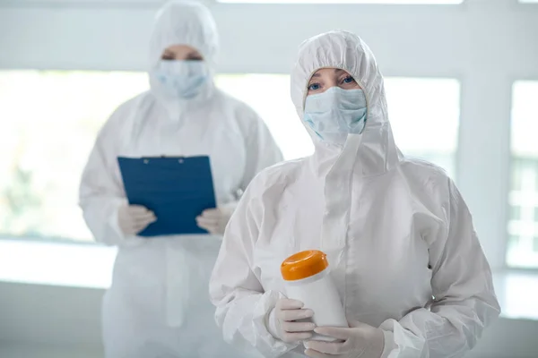 Медичний працівник в захисному одязі тримає пляшку для дезінфекції серветок, її колега стоїть позаду з папкою кліпу — стокове фото