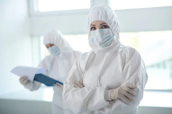 Жінка-медик у захисному одязі, що стоїть зі складеними руками, її колега стоїть позаду з папкою кліпу — стокове фото