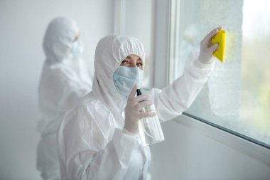 Koruyucu giysiler ve tıbbi maskeler giyen sağlık görevlileri pencere camlarını temizleyip dezenfekte ediyorlar.