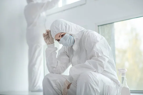 Trött läkare i skyddskläder och medicinsk mask sitter bredvid fönstret, hennes college arbetar bakom — Stockfoto