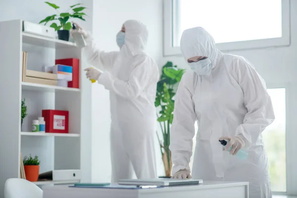 Medical worker in protective clothing and medical mask disinfecting table, another worker standing behind