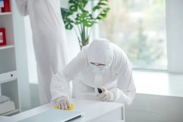 Medical worker in protective clothing and medical mask disinfecting table, another worker standing behind