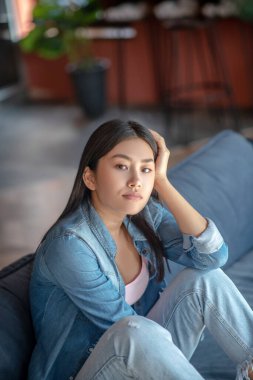 Koyu renk saçlı, dizleri katlanmış, koltukta oturan, düşünen, bir elinde dinlenen bir kadın.