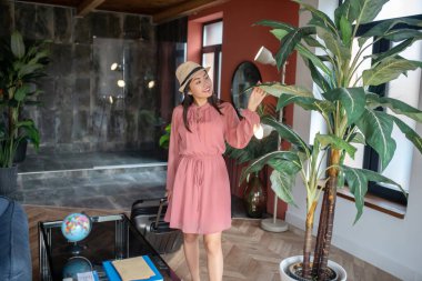 Hasır şapkalı heyecanlı bir kadın bavulunu çekiyor, ev bitkisinin yapraklarına dokunuyor.