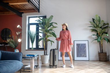 Hasır şapkalı, bavul sapını tutan, gülümseyen, heyecanlı siyah saçlı kadın.