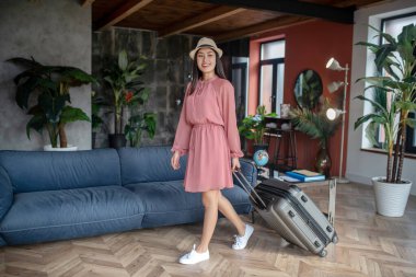 Hasır şapkalı, bavulunu çeken heyecanlı siyah saçlı kadın.