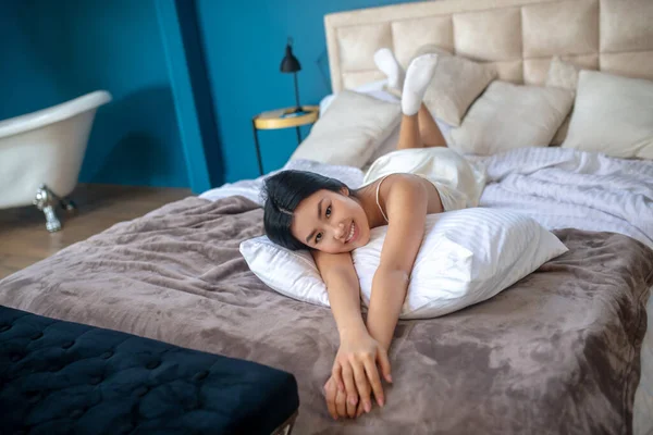 Молода брюнетка лежить на ліжку в нічній сукні, насолоджуючись ранком, відпочиваючи головою на плечі — стокове фото