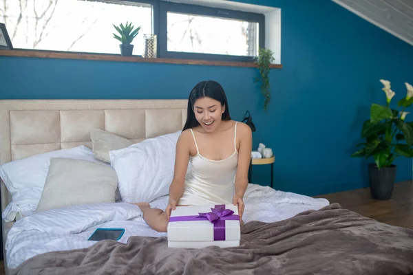 Joven morena en vestido de noche sentada en la cama, sonriendo, mirando a la caja actual con cinta púrpura — Foto de Stock