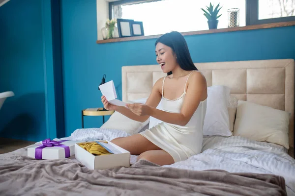 Młoda brunetka w nocnej sukience siedzi na łóżku z otwartym pudełkiem prezentów, czyta niektóre gazety, patrząc zaskoczony — Zdjęcie stockowe