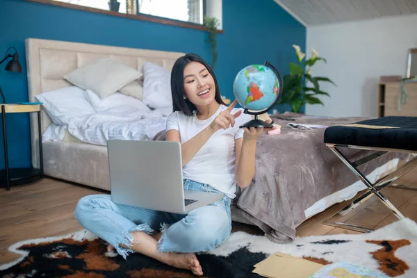 Młoda brunetka siedząca na dywanie z laptopem na kolanach, trzymająca kulę ziemską, wybierająca dokąd iść, uśmiechnięta — Zdjęcie stockowe