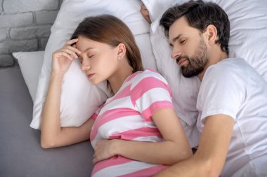 Koyu saçlı sakallı adam uyurken karısına sarılıyor.