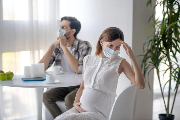 Mujer joven con una máscara facial y su marido sentado en la cocina y sintiéndose preocupado — Foto de Stock