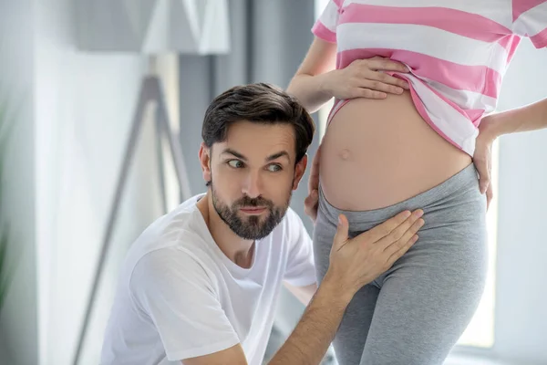 Ung skäggig man sätter örat nära sin gravida fru buken — Stockfoto