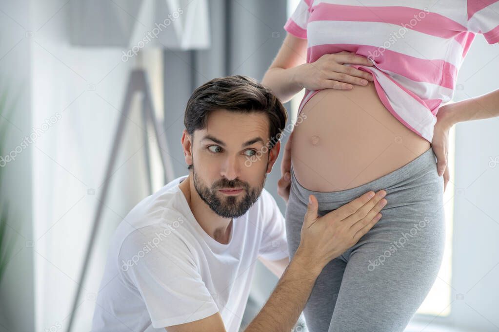 Young bearded man putting his ear close to his pregnant wifes abdomen