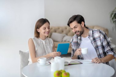 Beyaz elbiseli hoş bir genç kadın elinde tabletle kocasına bir şey gösteriyor.