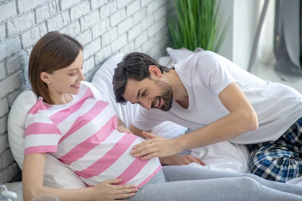 Homem de cabelos escuros sorrindo e tocando abdômen de sua esposa grávida — Fotografia de Stock