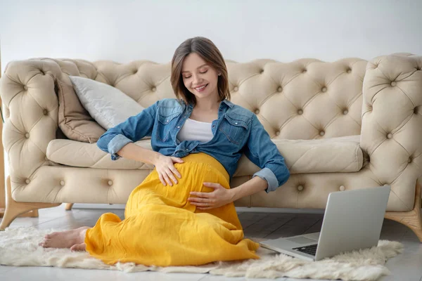 Junge schwangere Frau in Jeansjacke berührt ihren Bauch zärtlich und lächelt — Stockfoto