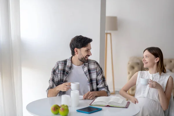 Sorrindo homem de cabelos escuros e sua esposa grávida sentada na cozinha e conversando — Fotografia de Stock