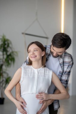 Koyu saçlı sakallı bir adam karısının karnına dokunuyor.