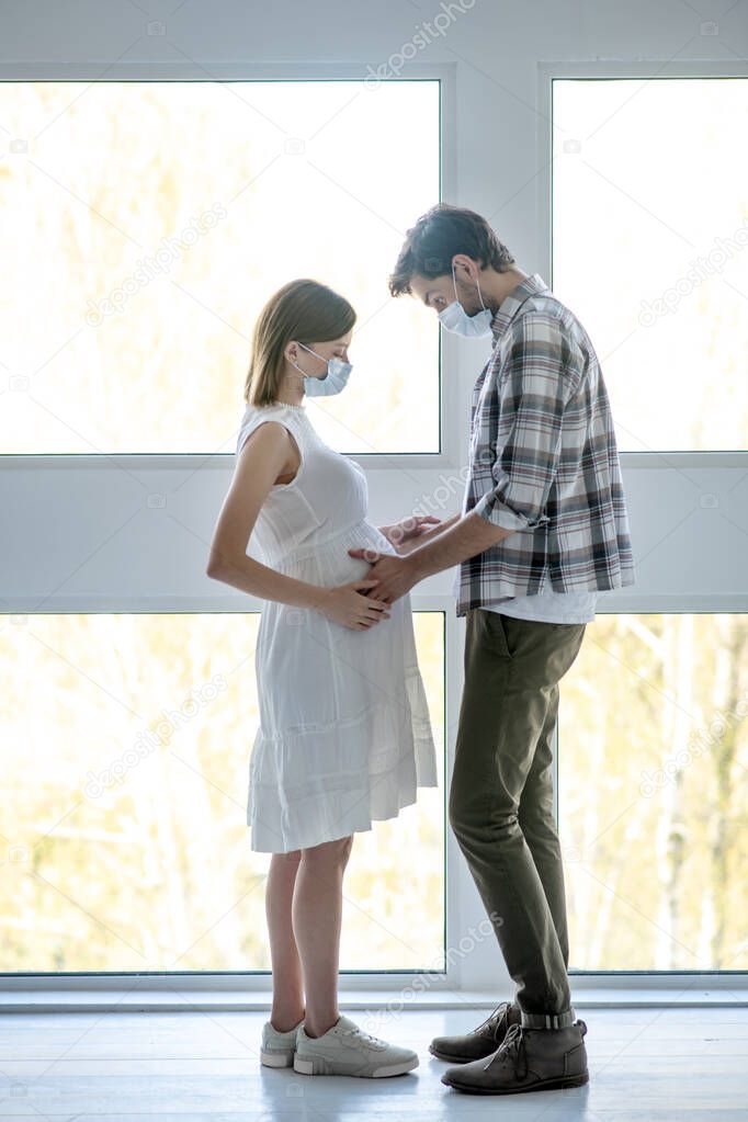 Tall man in a mask touching pregnant abdomen of his wife and feeling thrilled