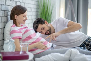 Koyu renk saçlı bir adam yatakta uzanıyor ve hamile karısının karnına dokunuyor.