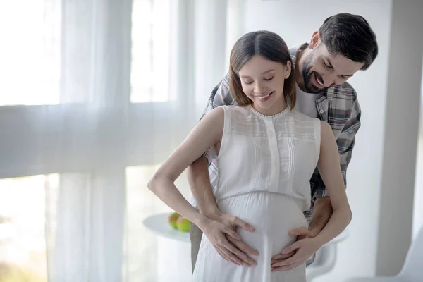 Mörkhårig skäggig man kramar sin gravida fru och ser glad ut — Stockfoto