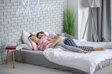 Koyu saçlı sakallı bir adam hamile karısına uyurken sarılıyor.