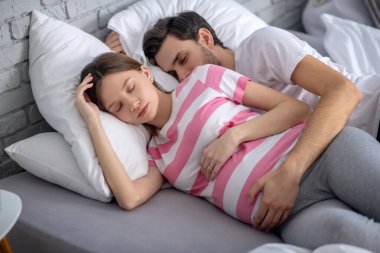 Koyu saçlı sakallı bir adam hamile karısına sarılıyor ve huzurlu görünüyor.