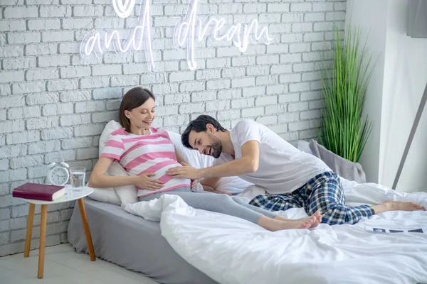 Mörkhårig man ligger i sängen och vidrör buken på sin gravida fru — Stockfoto