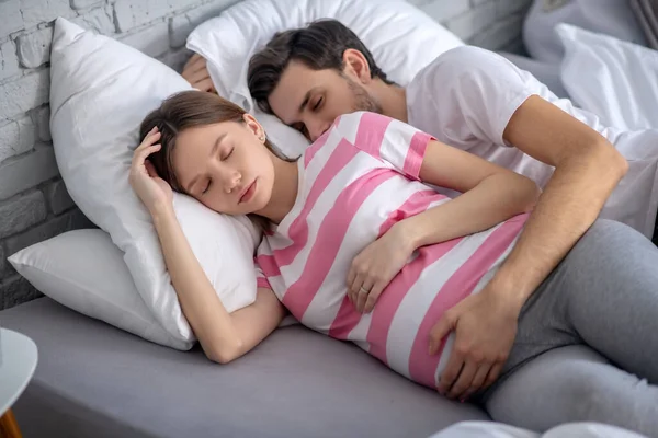 Темноволосый бородатый мужчина обнимает беременную жену во сне и выглядит умиротворенным. — стоковое фото