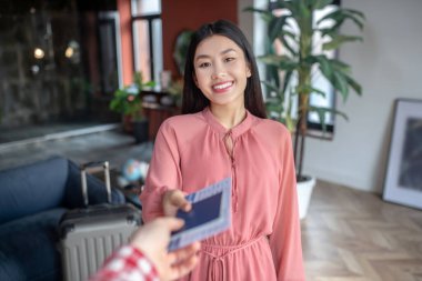 Neşeli siyah saçlı kadın, erkek ellerinden uçak bileti alıyor, gülümsüyor.