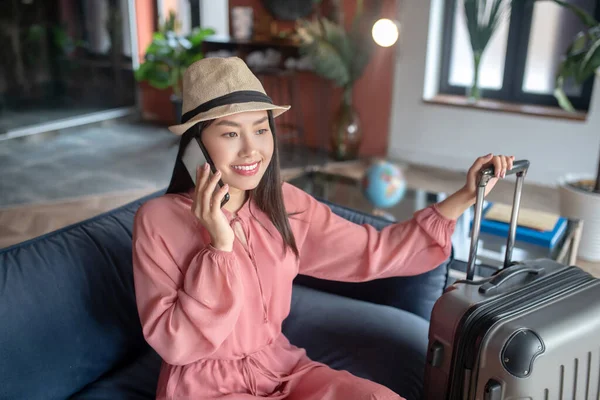 Темноволоса жінка в солом'яному капелюсі розмовляє на мобільному телефоні, сидить на дивані, тримає ручку валізи — стокове фото