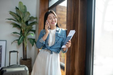 Koyu renk saçlı kadın kapı çerçevesine yaslanıyor, gülümsüyor, cep telefonuyla konuşuyor, uçak biletlerine bakıyor.