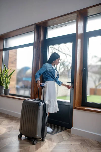Темноволоса жінка стоїть з валізою, відчиняючи скляні двері, залишаючи — стокове фото