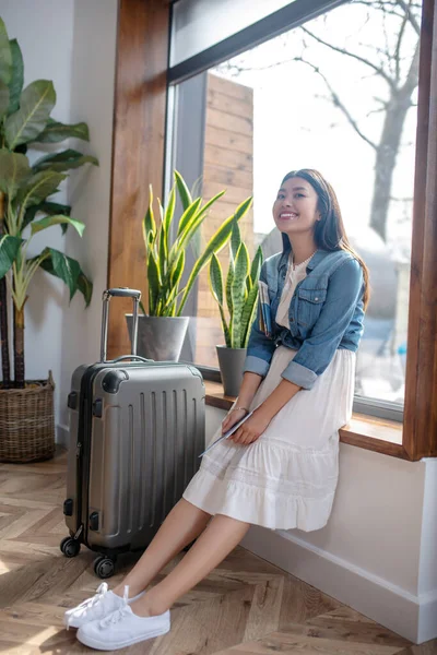 Dunkelhaarige Frau lehnt lächelnd am Fenster, ihr Koffer steht neben ihr — Stockfoto