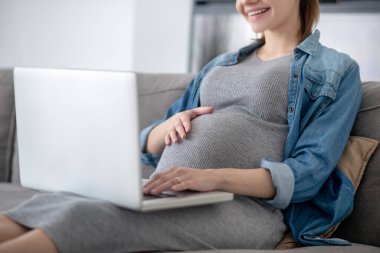 Elinde dizüstü bilgisayar tutan kot ceketli hamile bir kadın.