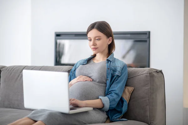 Junge schwangere Frau in Jeansjacke schaut sich online etwas an — Stockfoto