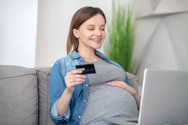 若いです妊娠中の女性でジーンズジャケットクレジットカードを保持し、オンラインショッピングを行う — ストック写真