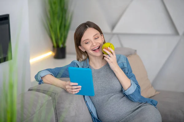 Ung gravid kvinna i jeans jacka sitter på soffan och äter äpple — Stockfoto