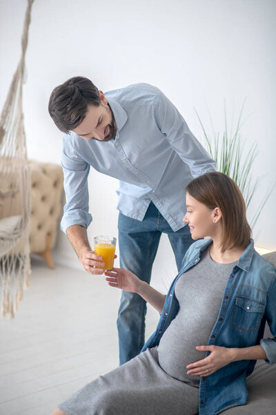 Темноволосый мужчина дарит стакан апельсинового сока своей беременной жене и улыбается
