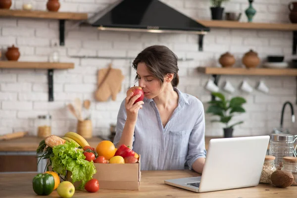 Mädchen in grauer Homewear sitzt am Tisch und riecht einen roten Apfel — Stockfoto